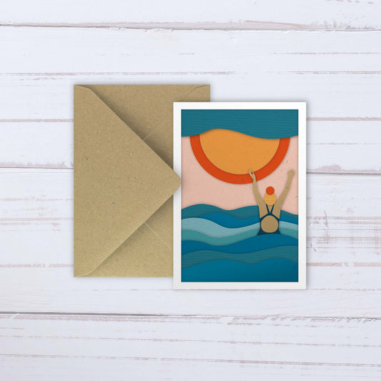 Wild sea swimming portrait A6 card featuring orange & yellow sun, blue waves and female with hands up in joy. Layered paper texture style. Brown C6 envelope